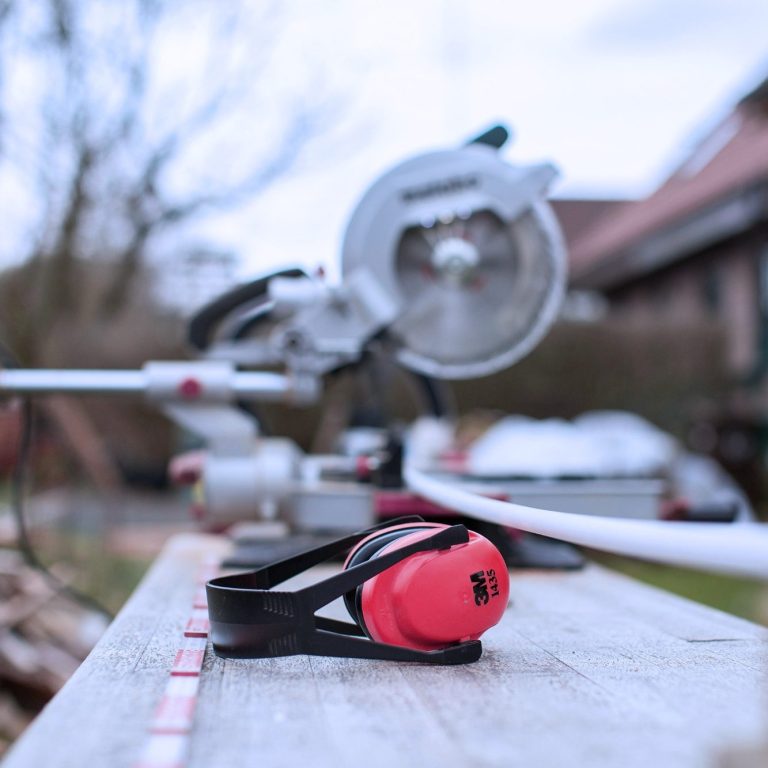 Protecțiile auditive roșii stau pe o suprafață de lemn lângă un ferăstrău electric.